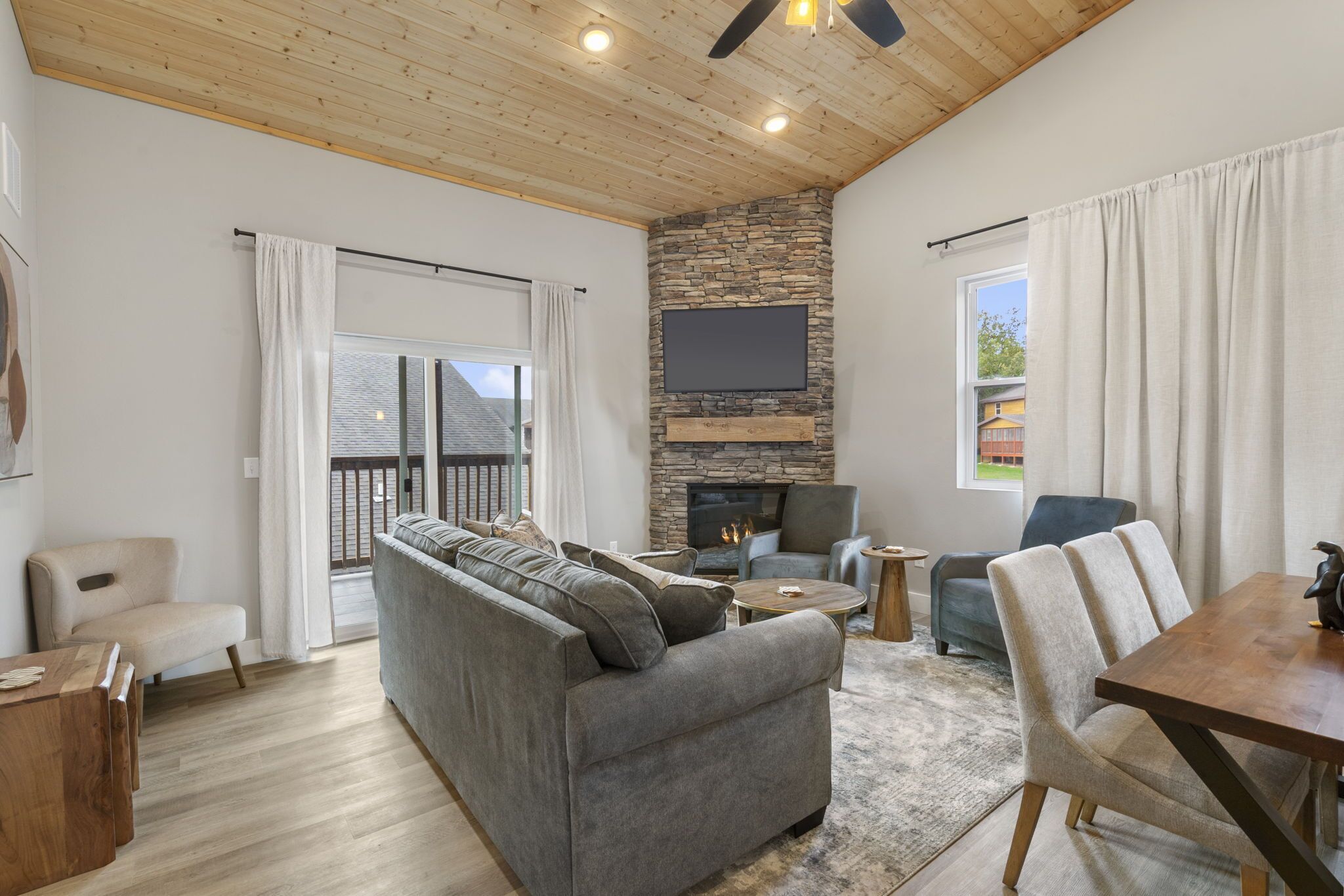 A spacious living room with a stone fireplace, a large flat-screen TV, and a wooden ceiling.