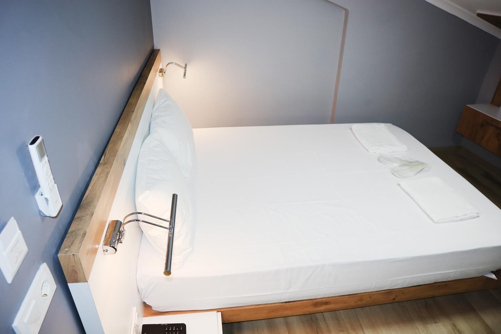 Una cama bien tendida, con un colchón y almohadas blancos, un cabecero de madera y una mesita de noche con una lámpara y un enchufe.