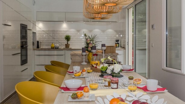 Una cocina moderna con azulejos blancos tipo metro, una mesa de comedor de madera con sillas amarillas, y una mesa llena de frutas y bebidas para el desayuno.