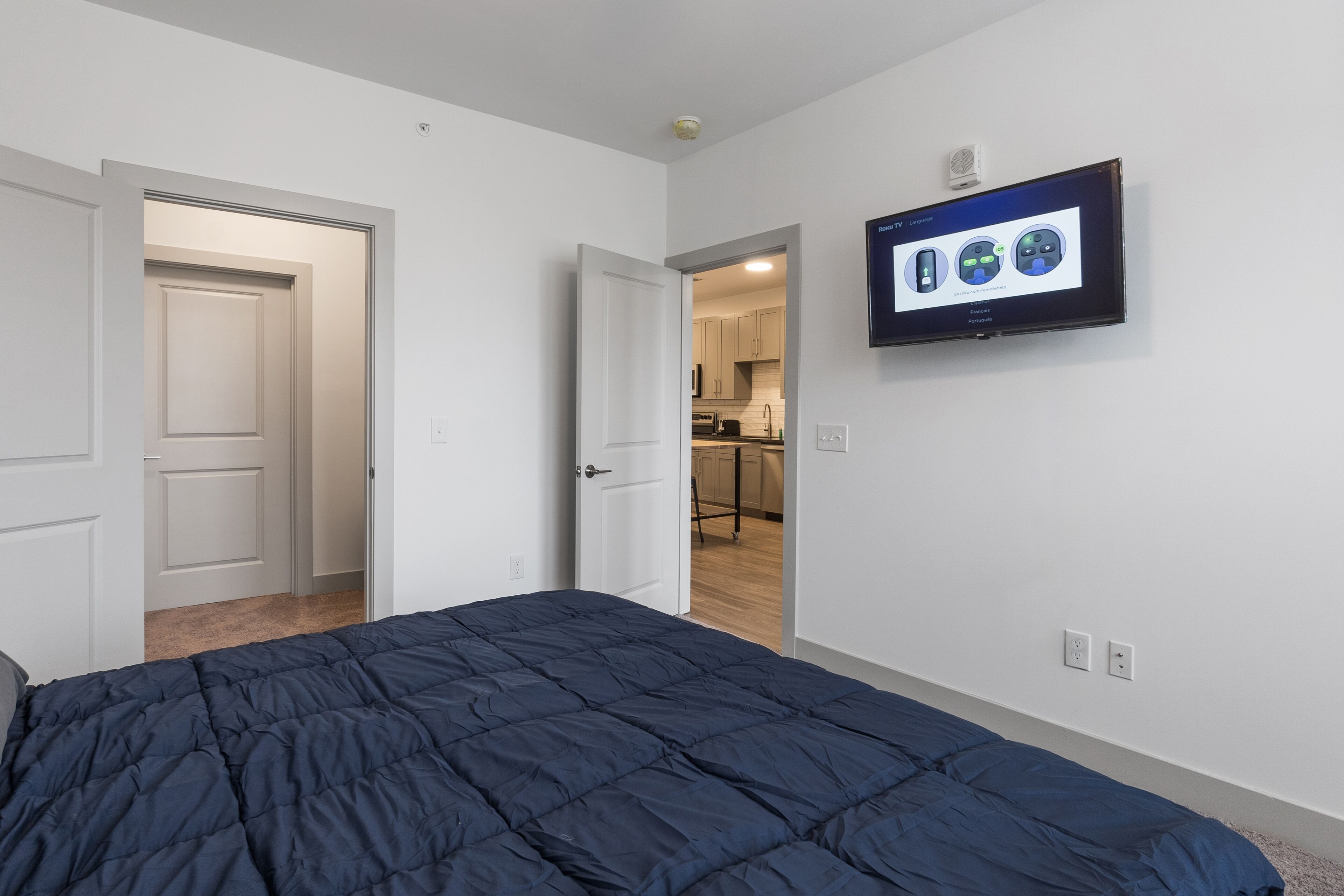 Un dormitorio con una cama, un televisor montado en la pared y una puerta abierta que da a la cocina.
