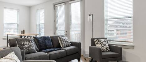 A modern living room with a grey sofa, armchairs, and a coffee table. There are large windows with blinds, a floor lamp, and a rug on the floor.