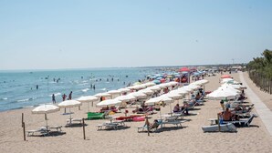 A beach with rows of sunbeds and umbrellas, a walkway along the shore, and people swimming in the water.