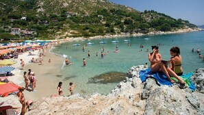 Ein Strand mit badenden und sonnenden Personen, einer felsigen Küstenlinie und einer Hügelkette mit Gebäuden im Hintergrund.