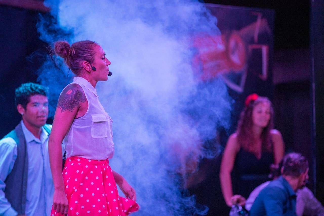 A person in a white vest and pink polka dot skirt standing in a room with a smoke effect in the background.