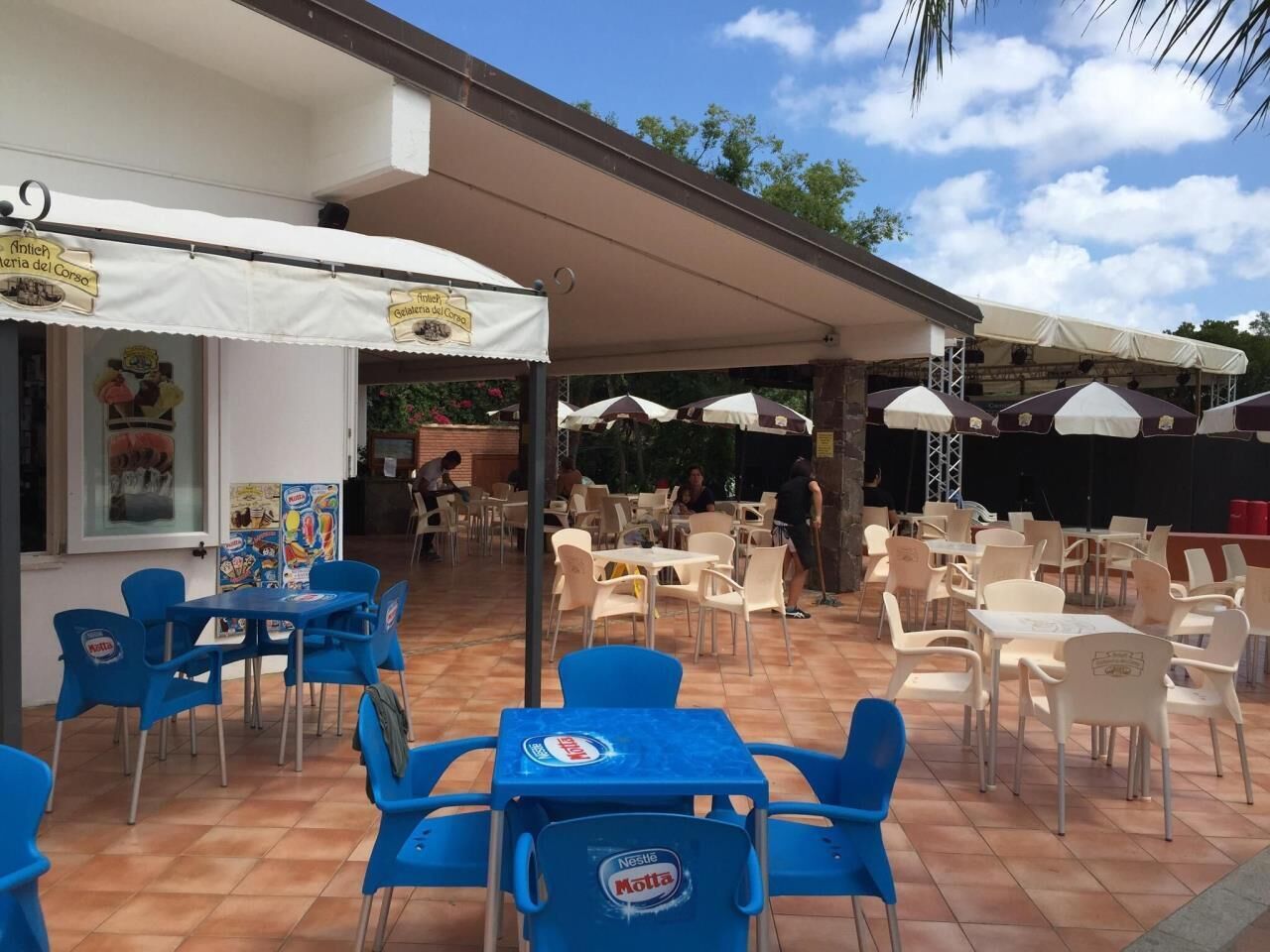 Ein Café im Freien mit Tischen und Stühlen, einige unter Sonnenschirmen, und ein Gebäude mit Schildern, die Speisen und Getränke anbieten.