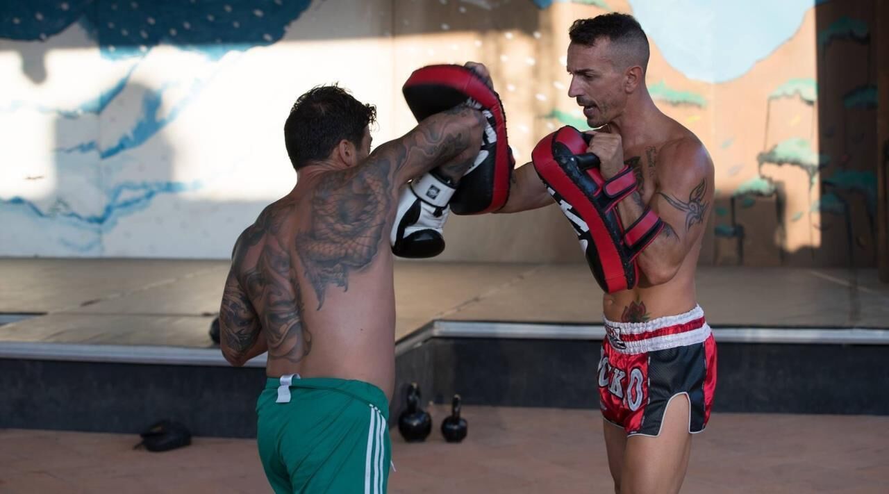 Two individuals in a gym setting, one wearing green shorts and the other in red and black shorts, practicing martial arts.