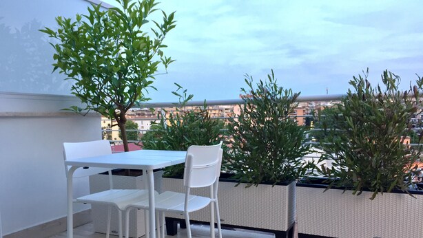 Eine Dachterrasse mit weißem Tisch und Stühlen, umgeben von Topfpflanzen und Blick auf eine Stadtlandschaft.