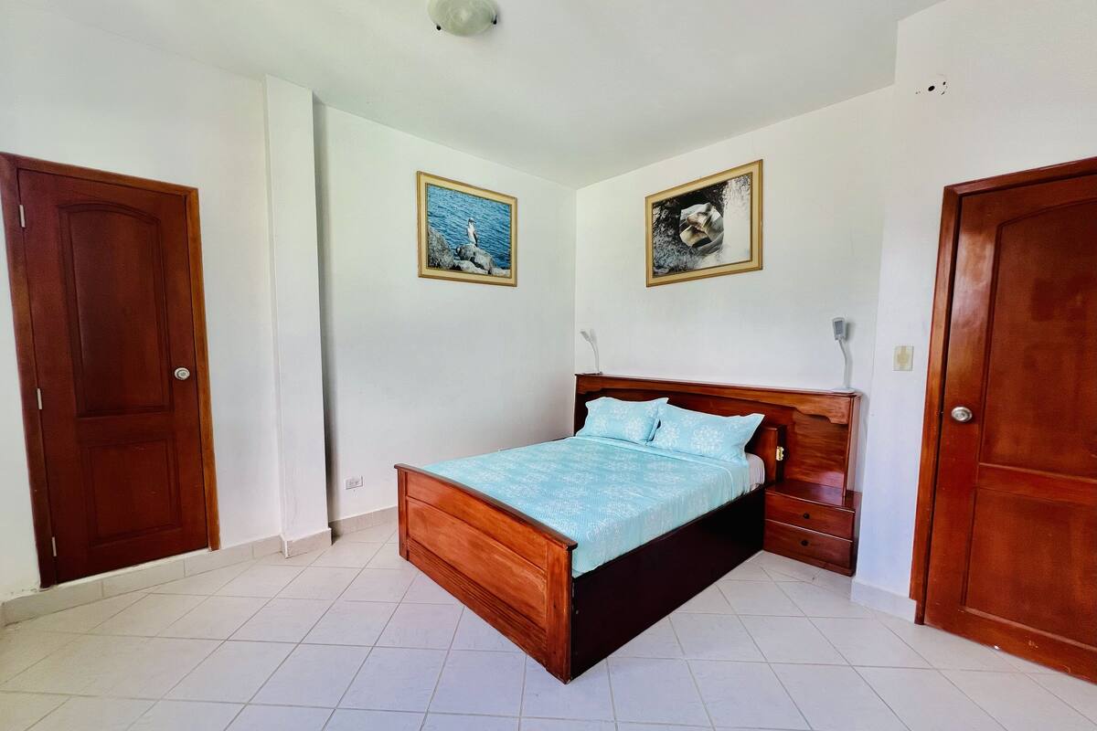 A bedroom with a wooden bed, white tiled floor, and two framed pictures on the wall.