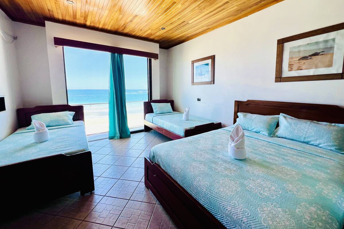 A hotel room with two beds, a wooden ceiling, a large window with a view of the beach, and a framed picture on the wall.