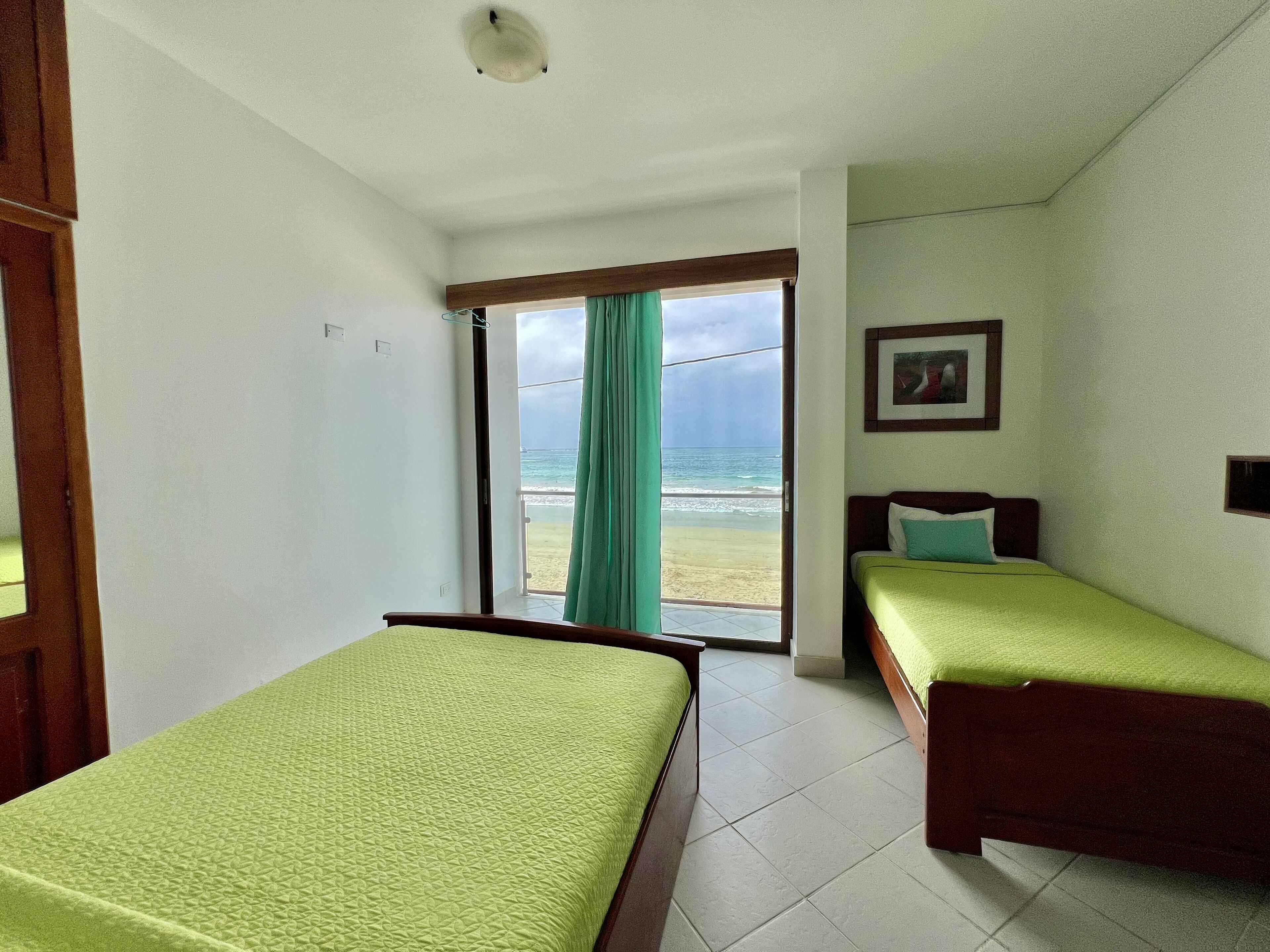 Une chambre avec deux lits, une porte donnant sur un balcon avec vue sur la plage, et un tableau encadré au mur.