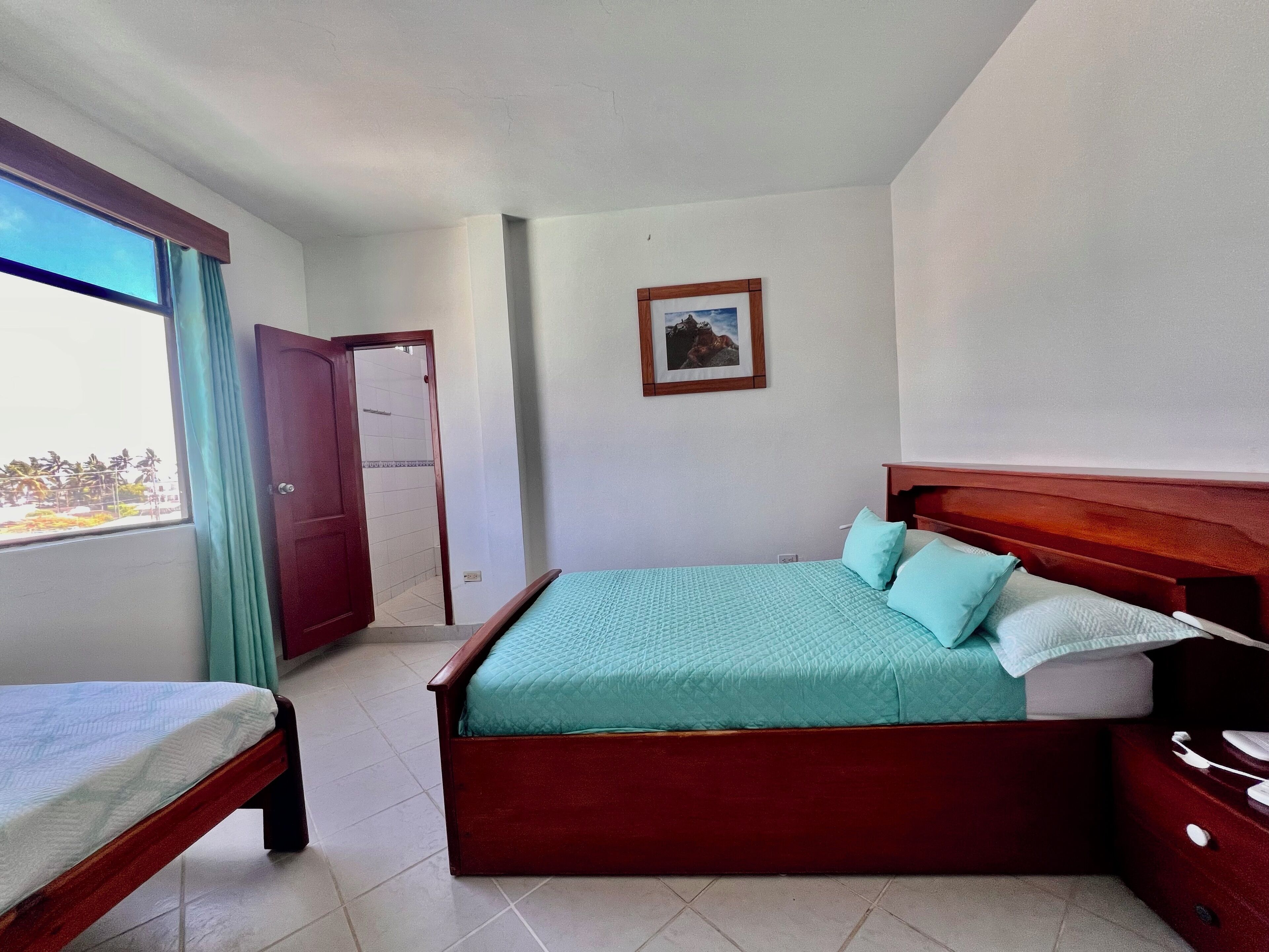 A bedroom with a bed, a bedside table, a window with curtains, and a door leading to a bathroom.