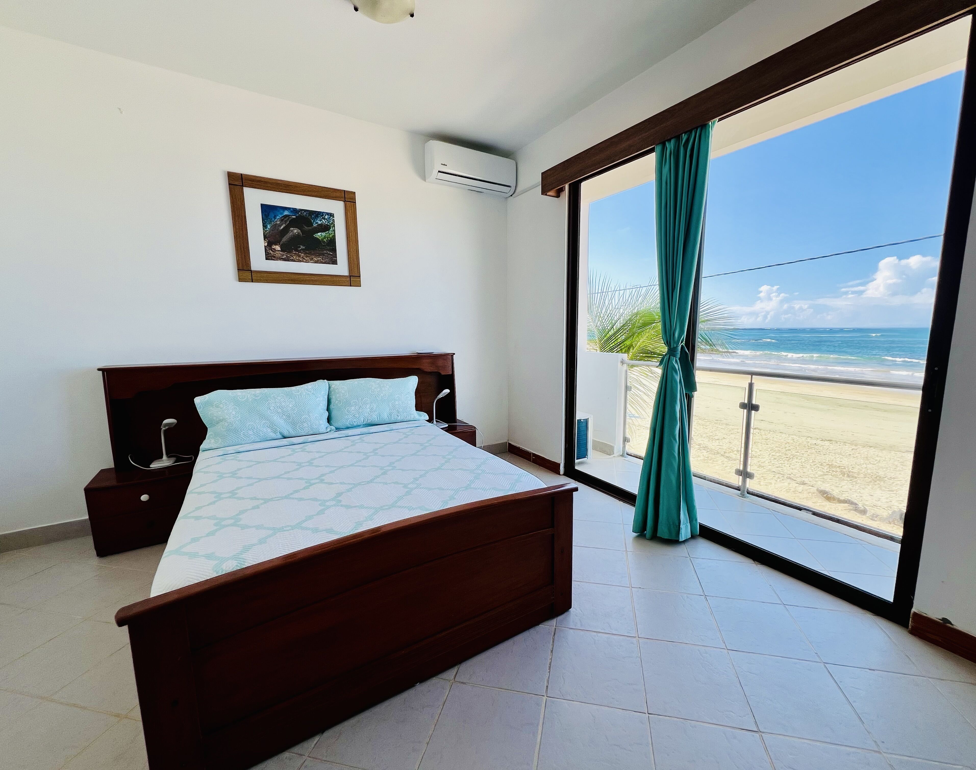 Camera da letto con un letto, un comodino, condizionatore d'aria a muro, un quadro incorniciato e un'ampia porta scorrevole in vetro che dà su una spiaggia.