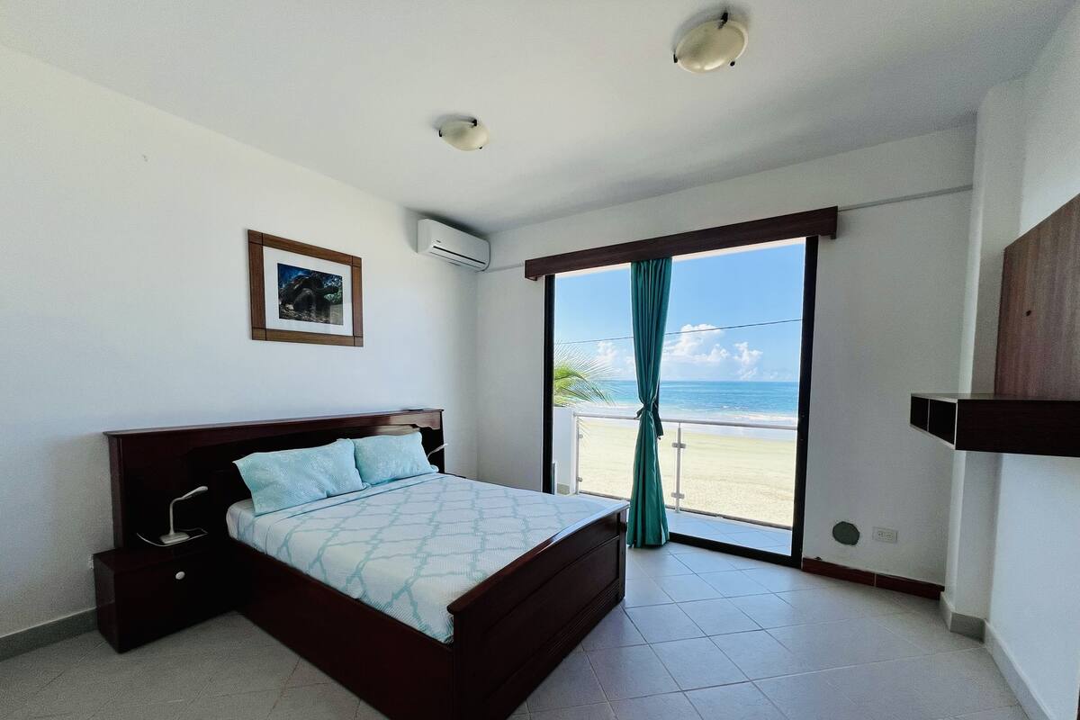 A bedroom with a bed, a picture on the wall, an air conditioning unit, and a view of the beach through a sliding door.