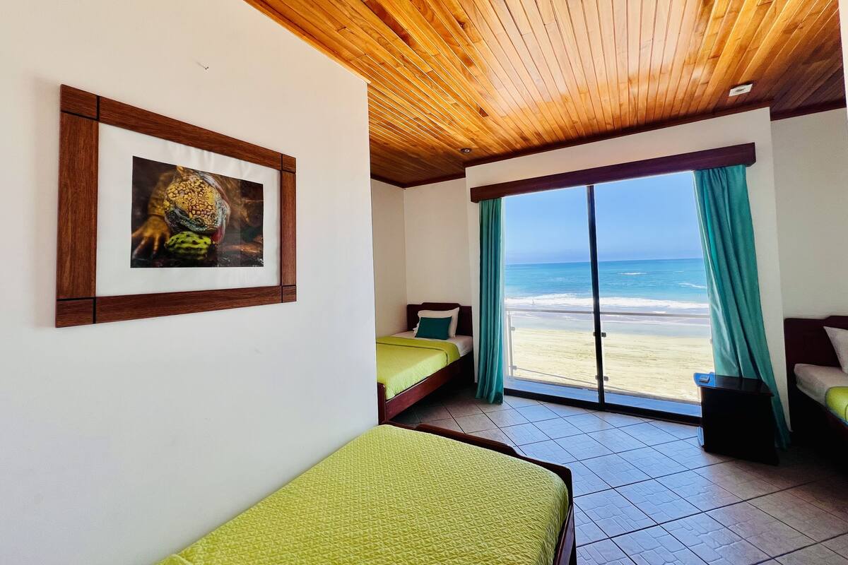 A room with a wooden ceiling, a bed with a green bedspread, a framed picture on the wall, and a large window with a view of the beach.