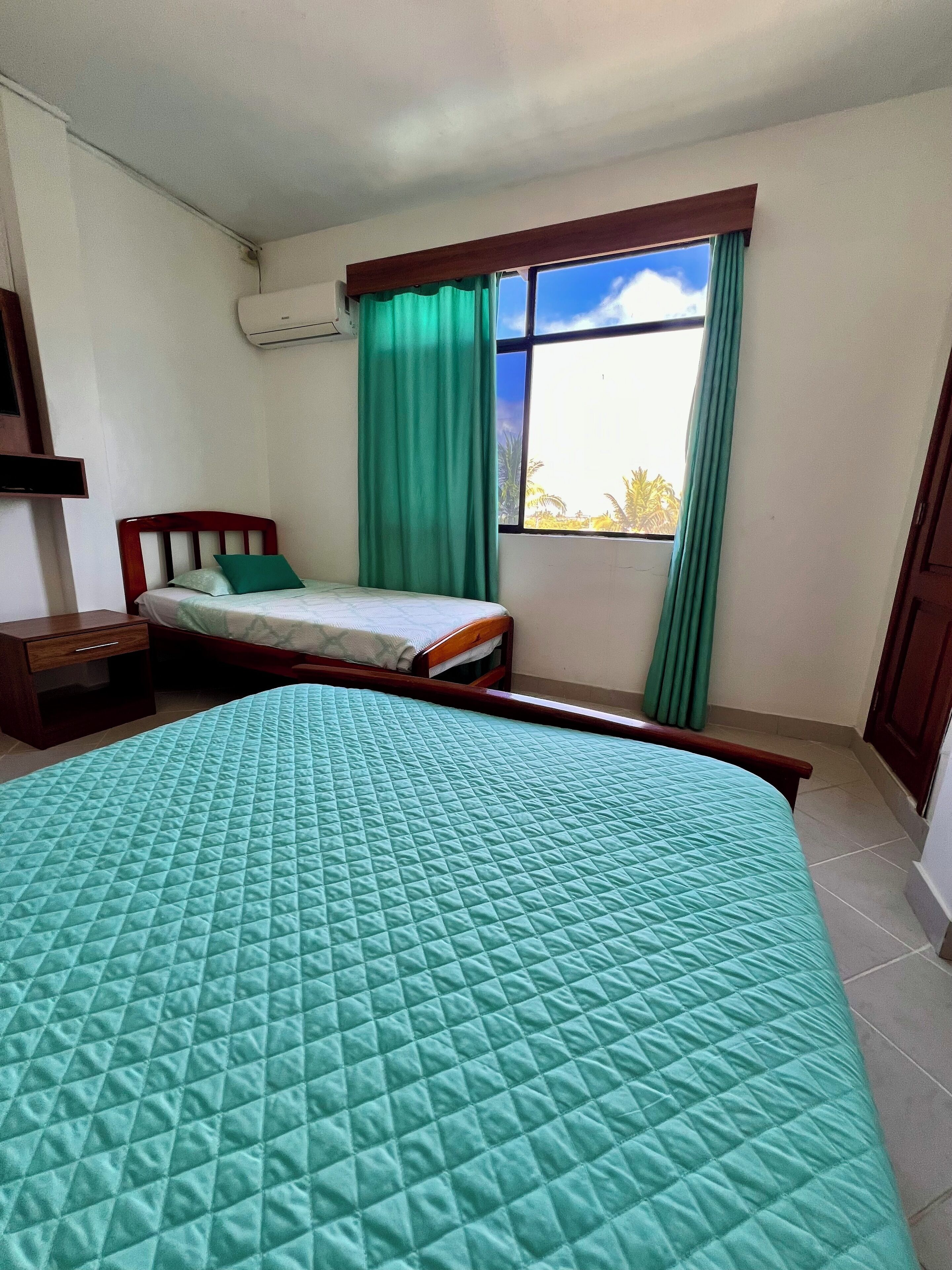 A bedroom with a large bed, a smaller bed, a nightstand, a window with curtains, and an air conditioning unit.