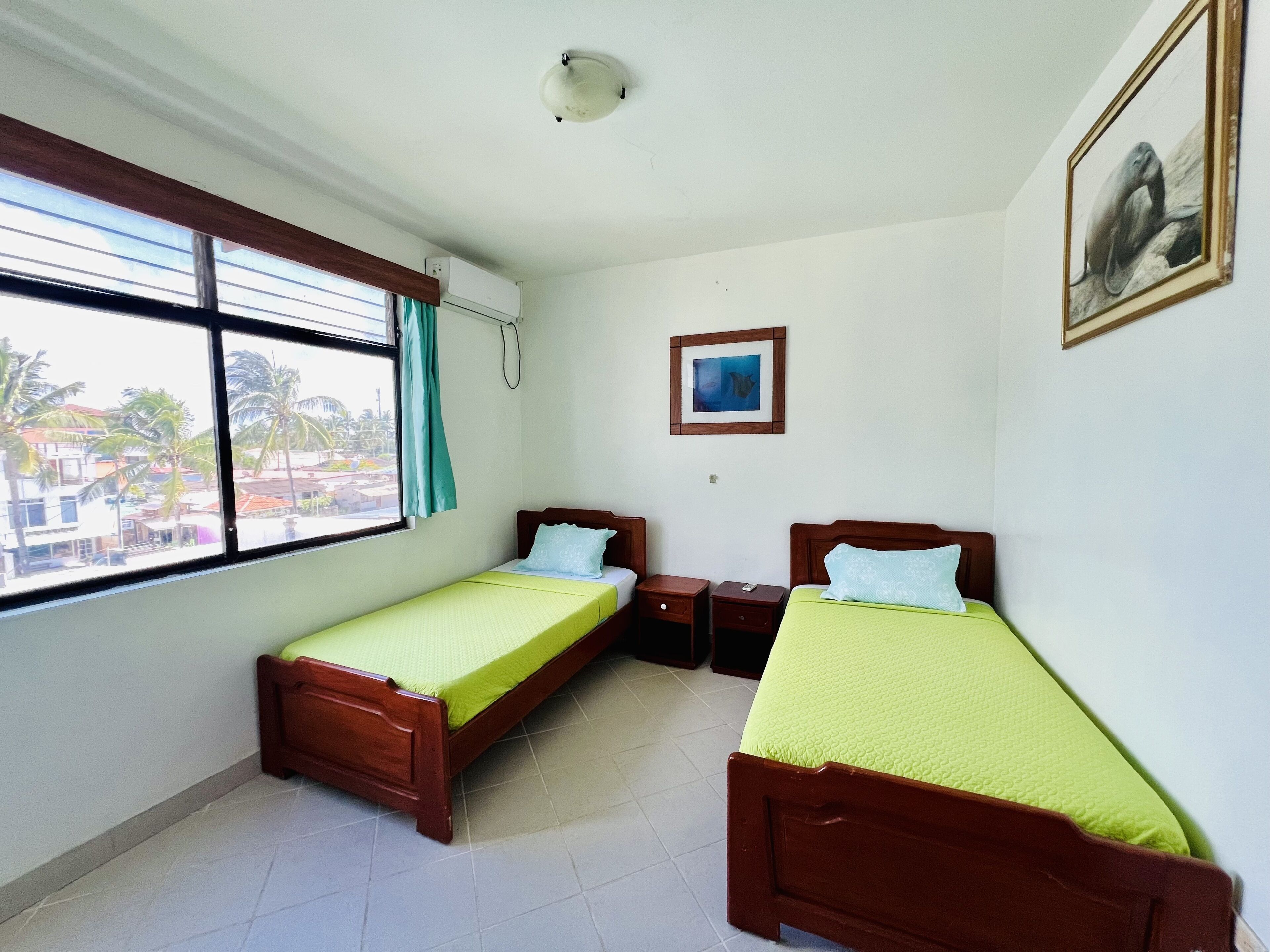 A room with two wooden beds, a window with curtains, a ceiling light, a framed picture on the wall, and a tiled floor.
