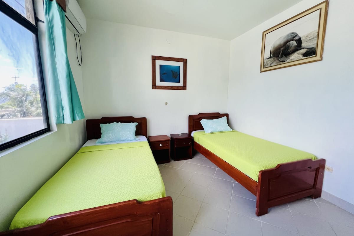 A room with two wooden beds, each with a green bedspread and a pillow. There is a framed picture on the wall and a window with a curtain.