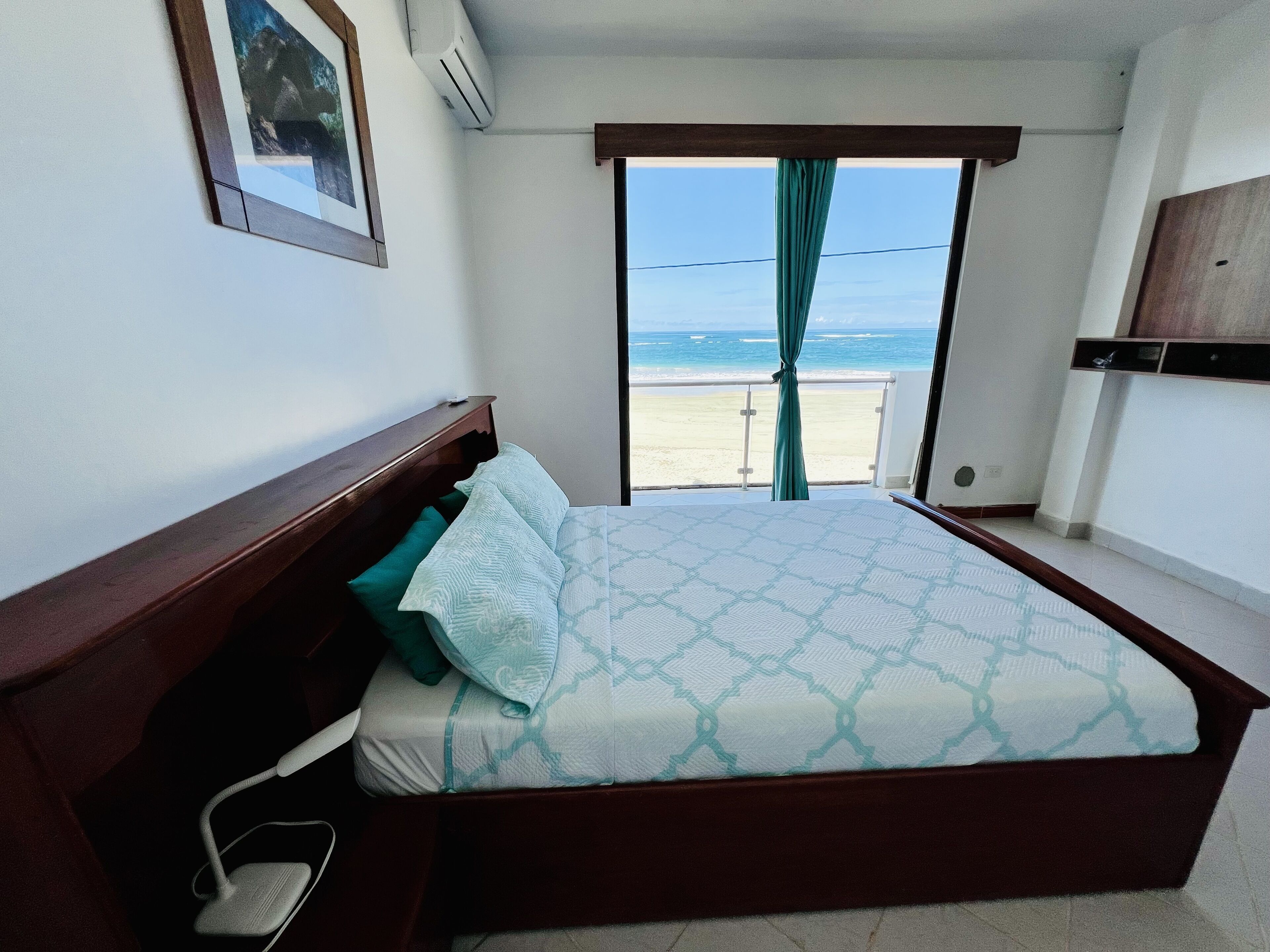 A bedroom with a large bed, a bedside table, a nightstand, a window with a view of the beach, and a mounted TV.