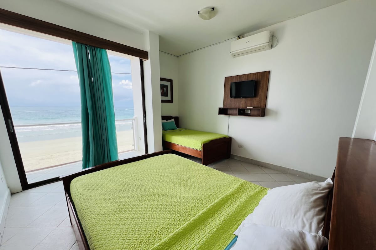A hotel room with a bed, a TV mounted on the wall, and a view of the beach through the window.