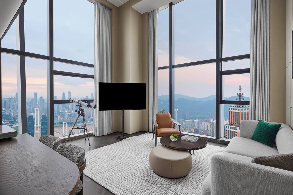 A modern hotel room with a large city view, a flat-screen TV, a telescope, and a comfortable seating area.