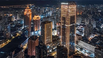 Une vue nocturne d’une ville, avec de nombreux gratte-ciel bien visibles, dont un bâtiment portant l’inscription « CONRAD ».