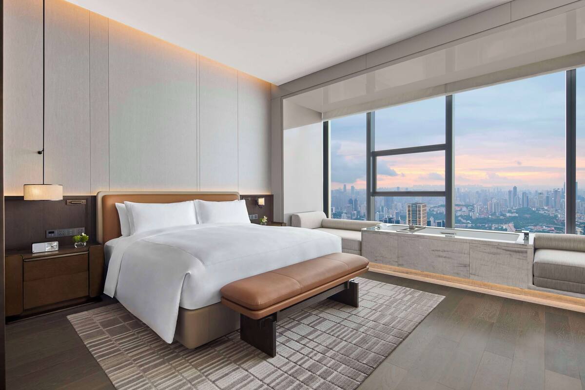 A modern hotel room with a large bed, a bench, a nightstand, a lamp, and a view of the city through large windows.