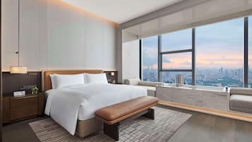 A modern hotel room with a large bed, a bench, a nightstand, a lamp, and a view of the city through large windows.