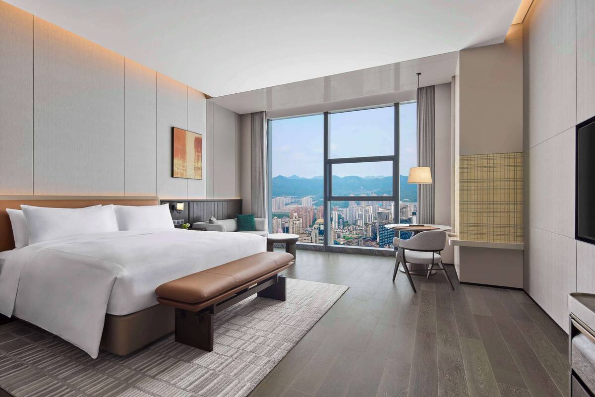 A modern hotel room with a large bed, a bench, a desk, and a chair. There is a large window with a city view.