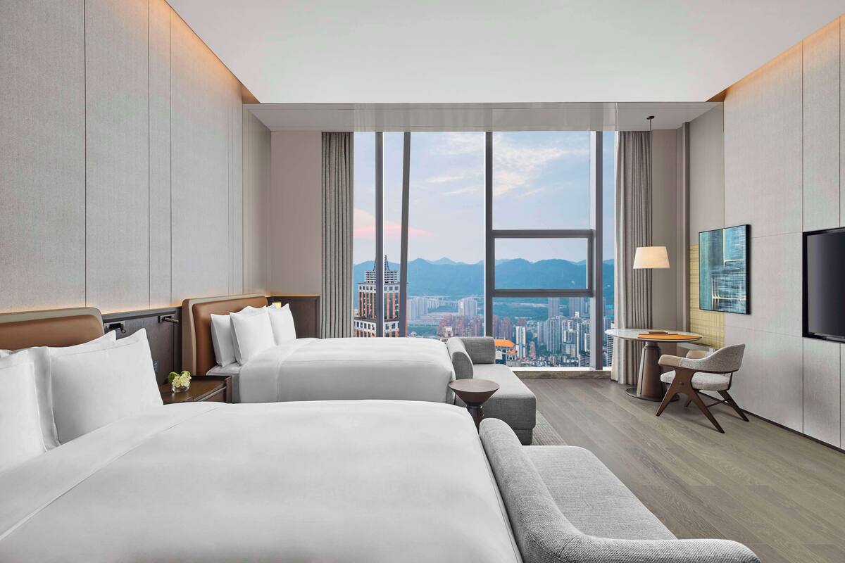 A hotel room with two beds, a desk, a chair, and a large window offering a city view.