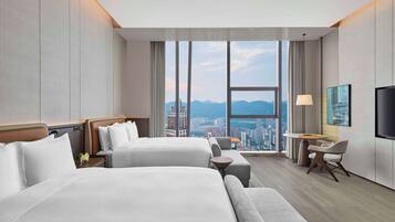 A hotel room with two beds, a desk, a chair, and a large window offering a city view.
