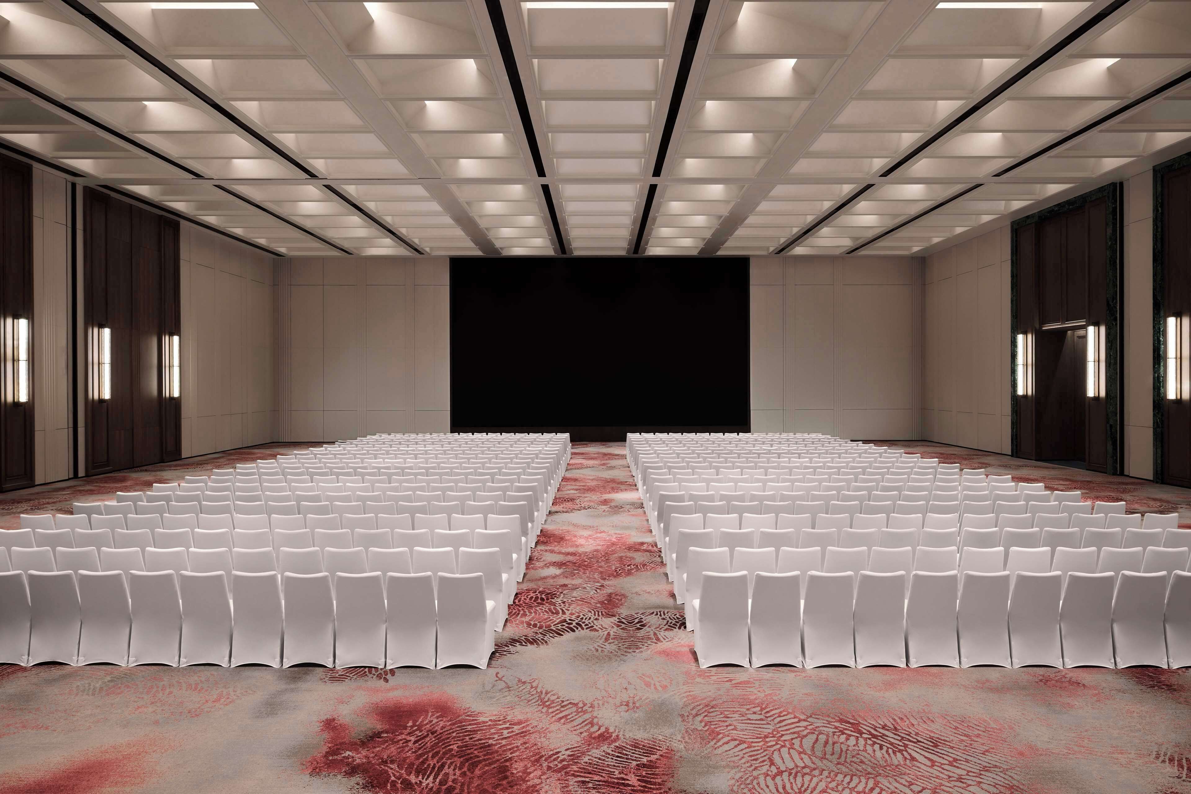 A conference room with rows of white chairs, a patterned carpet, and a dark screen at the front.