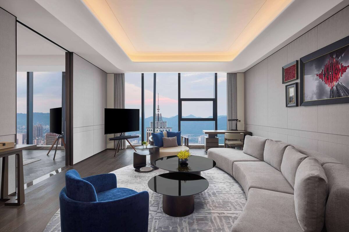 A modern living room with a sofa, a coffee table, and a TV. There is a large window with a view of a cityscape.