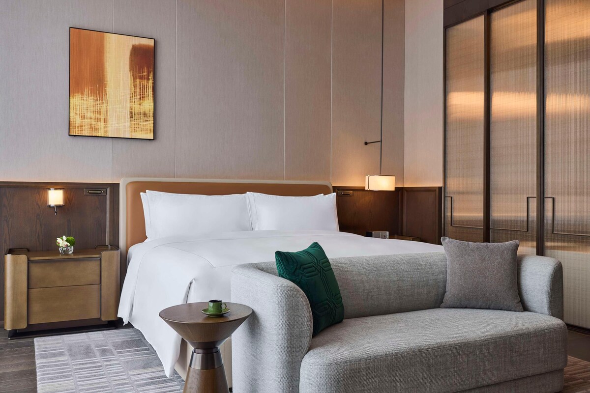 Una habitación de hotel con una cama grande, un sofá, una mesita de noche con lámpara y una obra de arte en la pared.