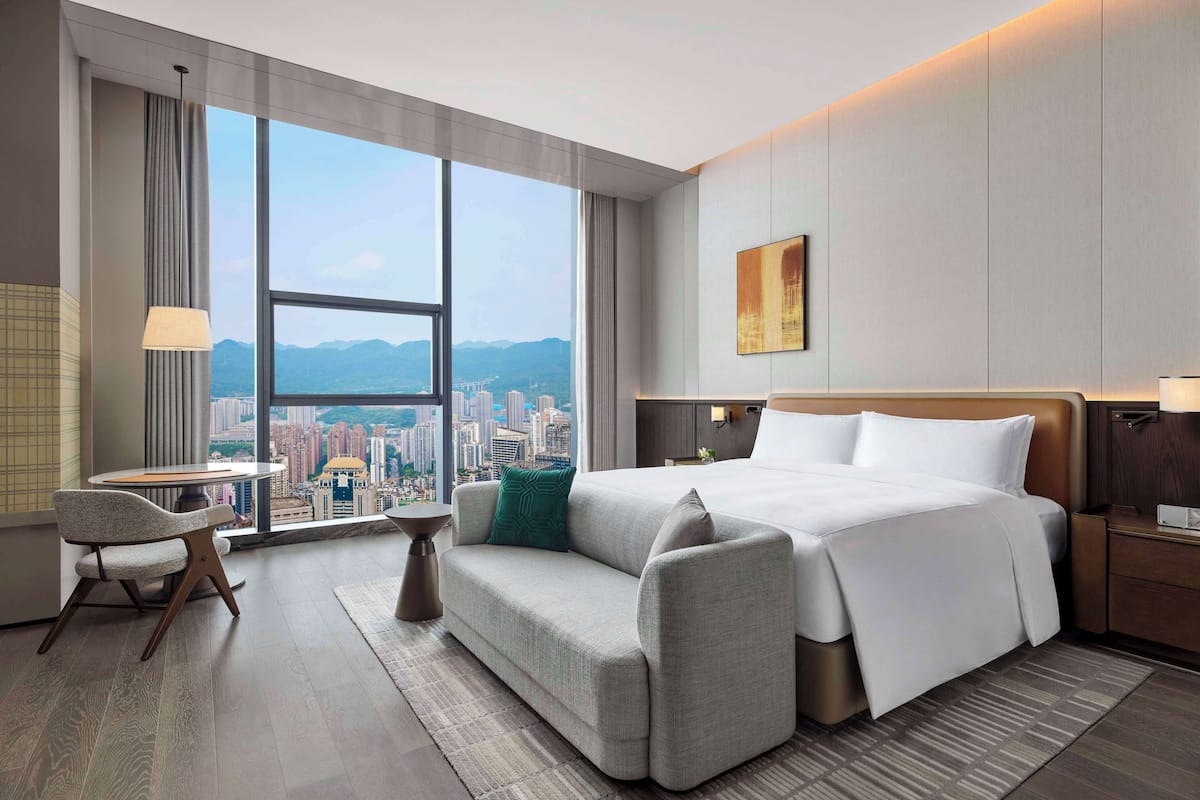 A modern hotel room with a large window offering a city view, a comfortable sofa, a neatly made bed, and a small table.