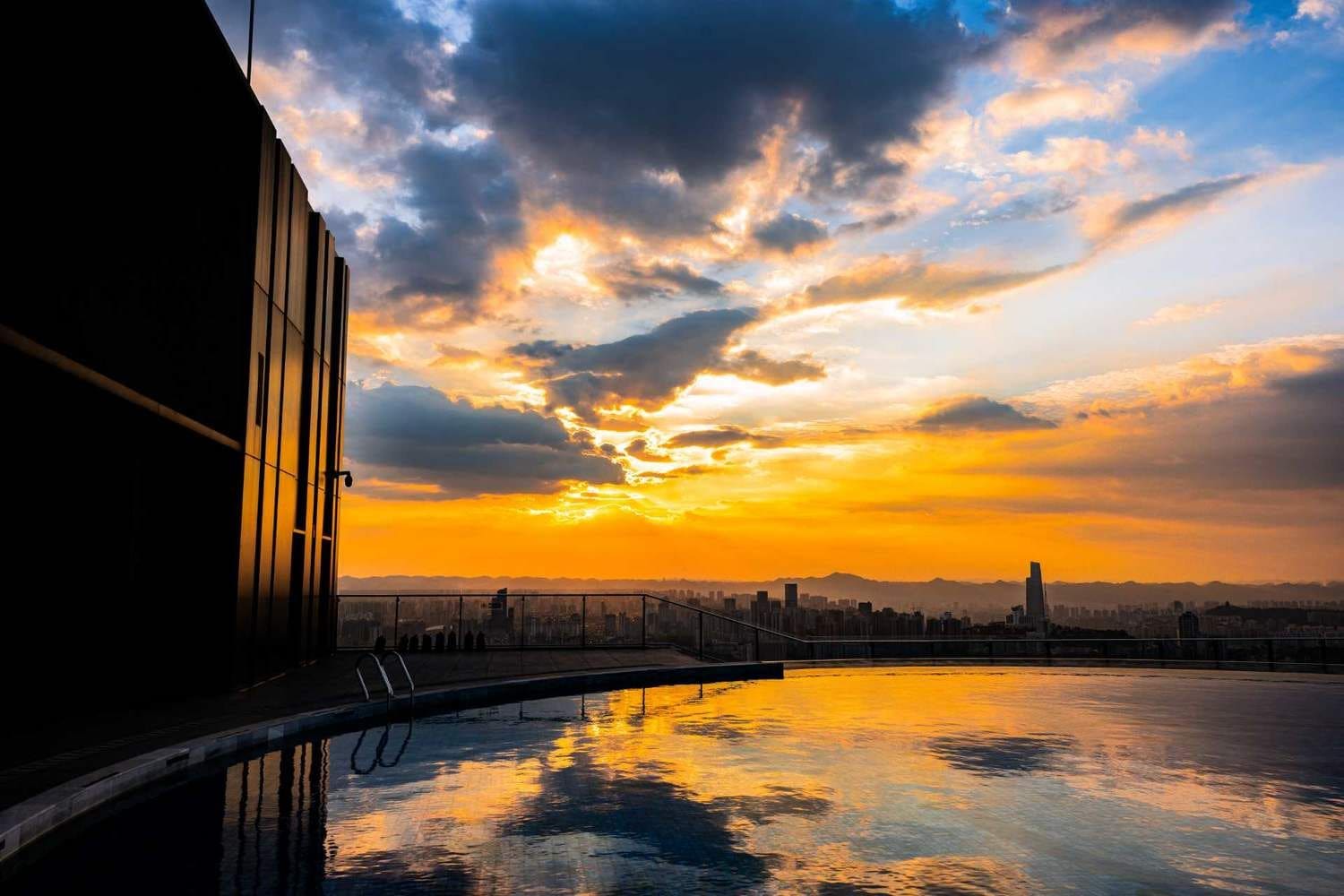 A sunset view from a rooftop terrace with a pool, overlooking a cityscape.