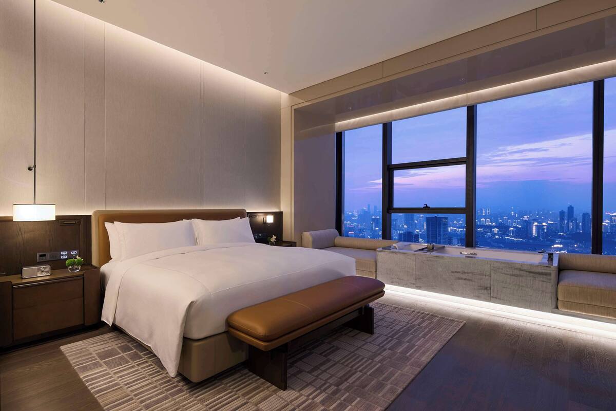 A modern hotel room with a large bed, a bench, a nightstand, and a view of the city at dusk.