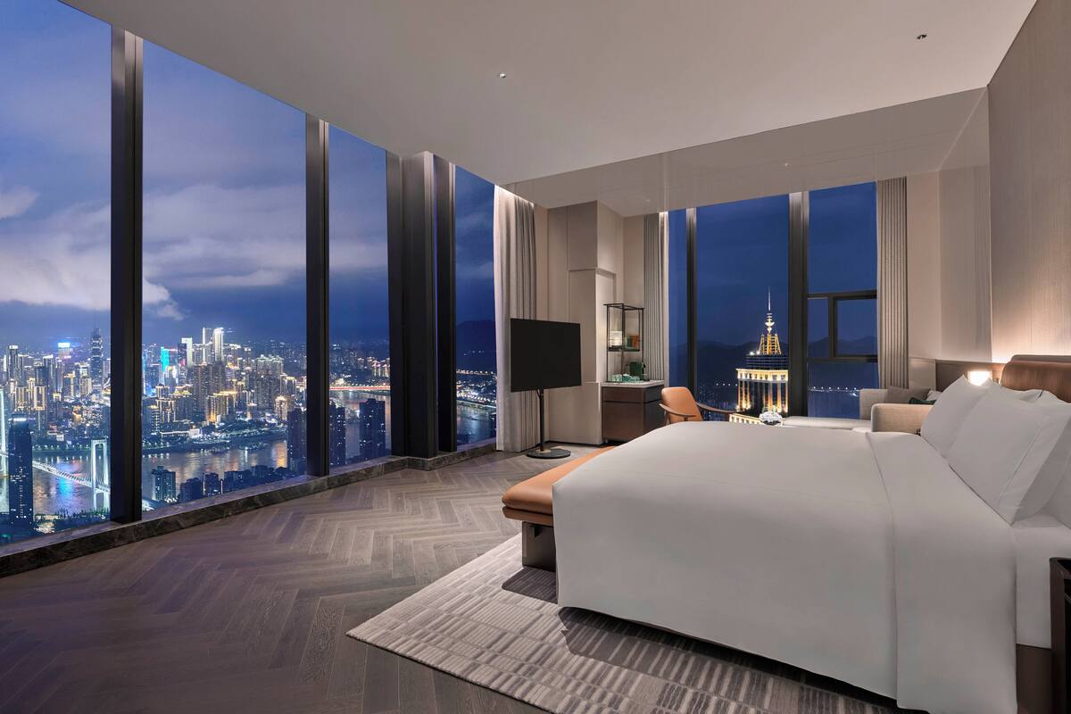 A modern hotel room with a large bed, a desk, and a chair. It features a city view through large windows.