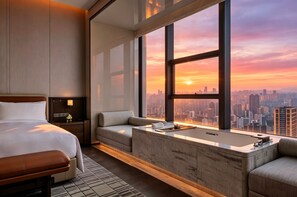 A modern hotel room with a large bed, a sofa, a bathtub, and a city view at sunset.