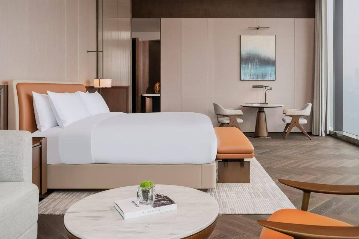 A modern hotel room with a large bed, a small desk, a round table, and a view of the city.
