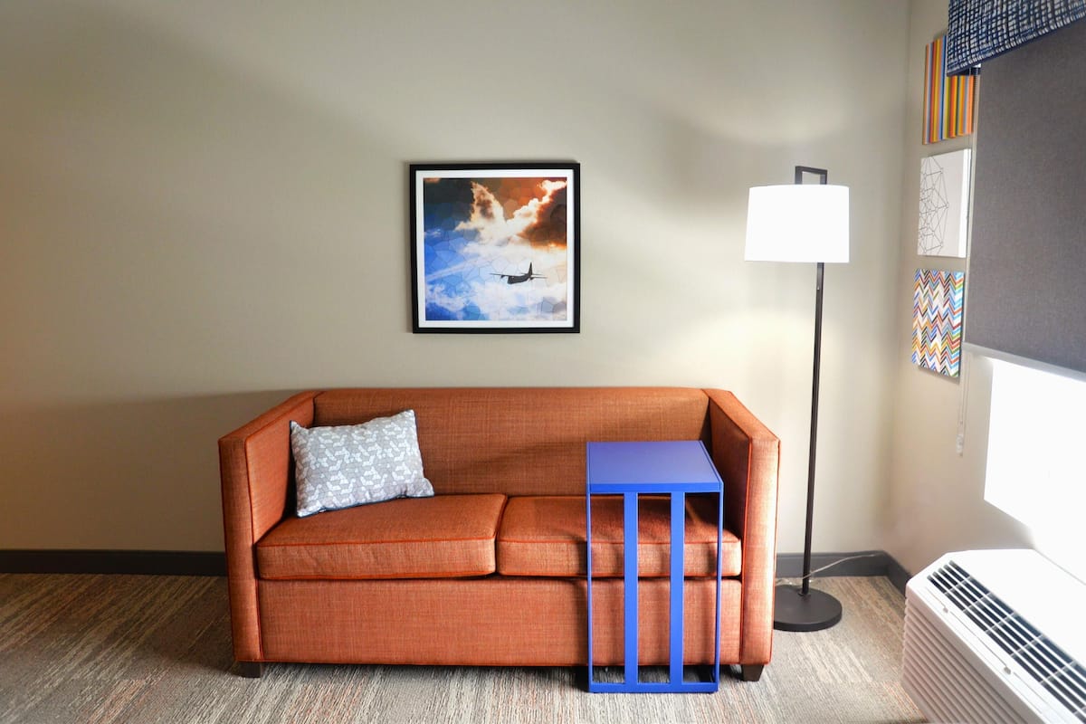 Een kamer met een oranje bank, een blauwe bijzettafel, een ingelijst beeld van een vliegtuig, een staande lamp en een muur met kleurrijke kunstwerken.