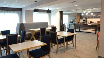 A modern dining area with wooden tables and blue chairs, a kitchen in the background, and a large window providing natural light.