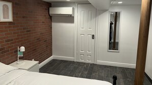 Een slaapkamer met een bakstenen muur, een witte deur met een keypad, een nachtkastje met een lamp en een bed met witte beddengoed.
