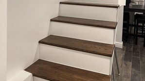 Een trap met houten treden en witte wanden, die leidt naar een keuken met donkere vloer.