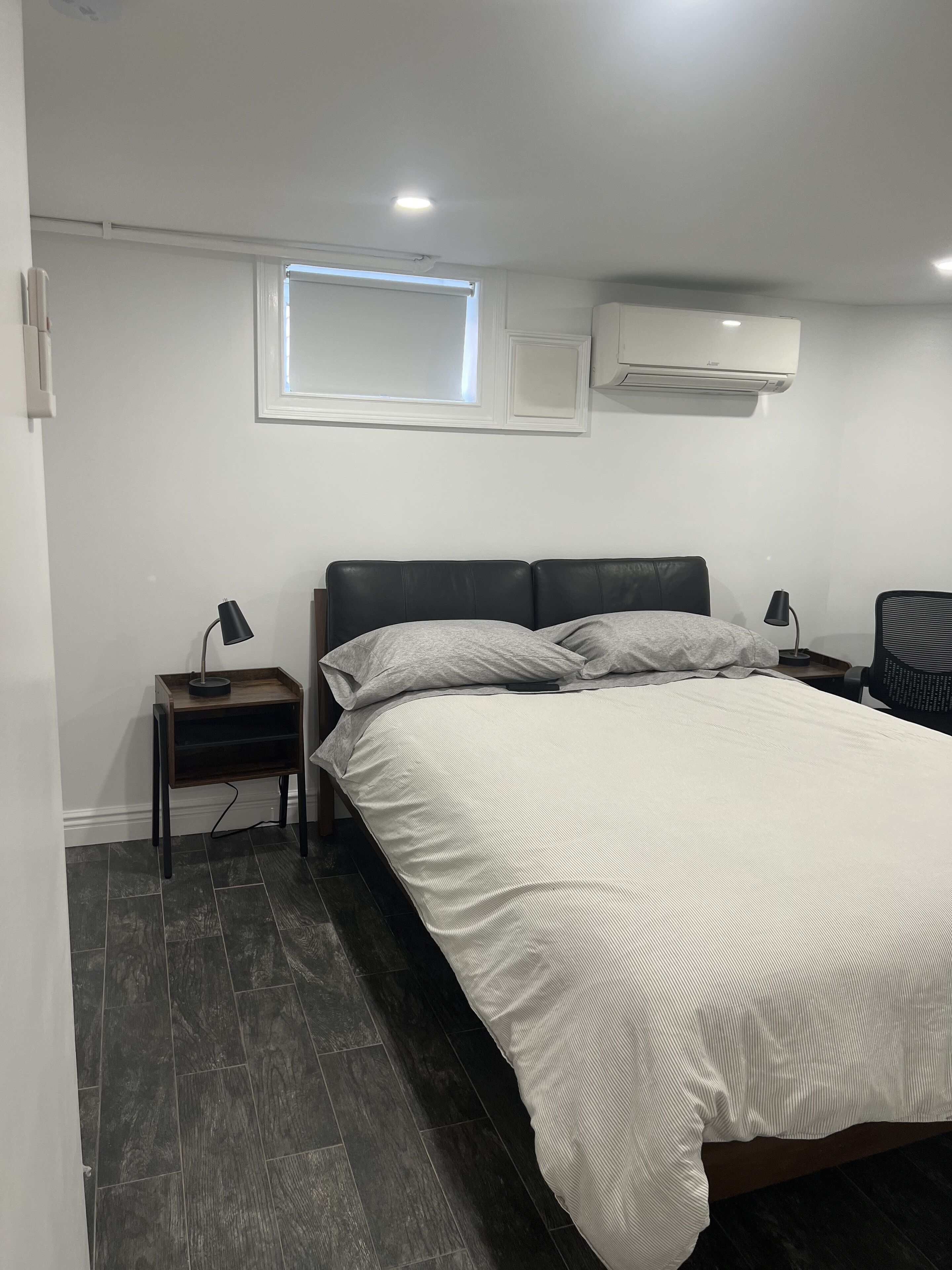 Un dormitorio moderno con una cama grande, una mesita de noche con lámpara, un estante pequeño y un equipo de aire acondicionado en la pared.