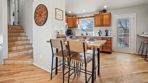 Una cocina con armarios de madera, una mesa de comedor con sillas, un reloj en la pared y una escalera que lleva a otra habitación.