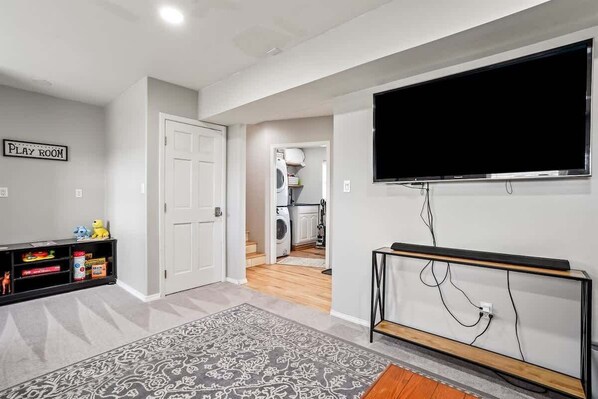 A playroom with a television, a washing machine, and a kitchen in the background.