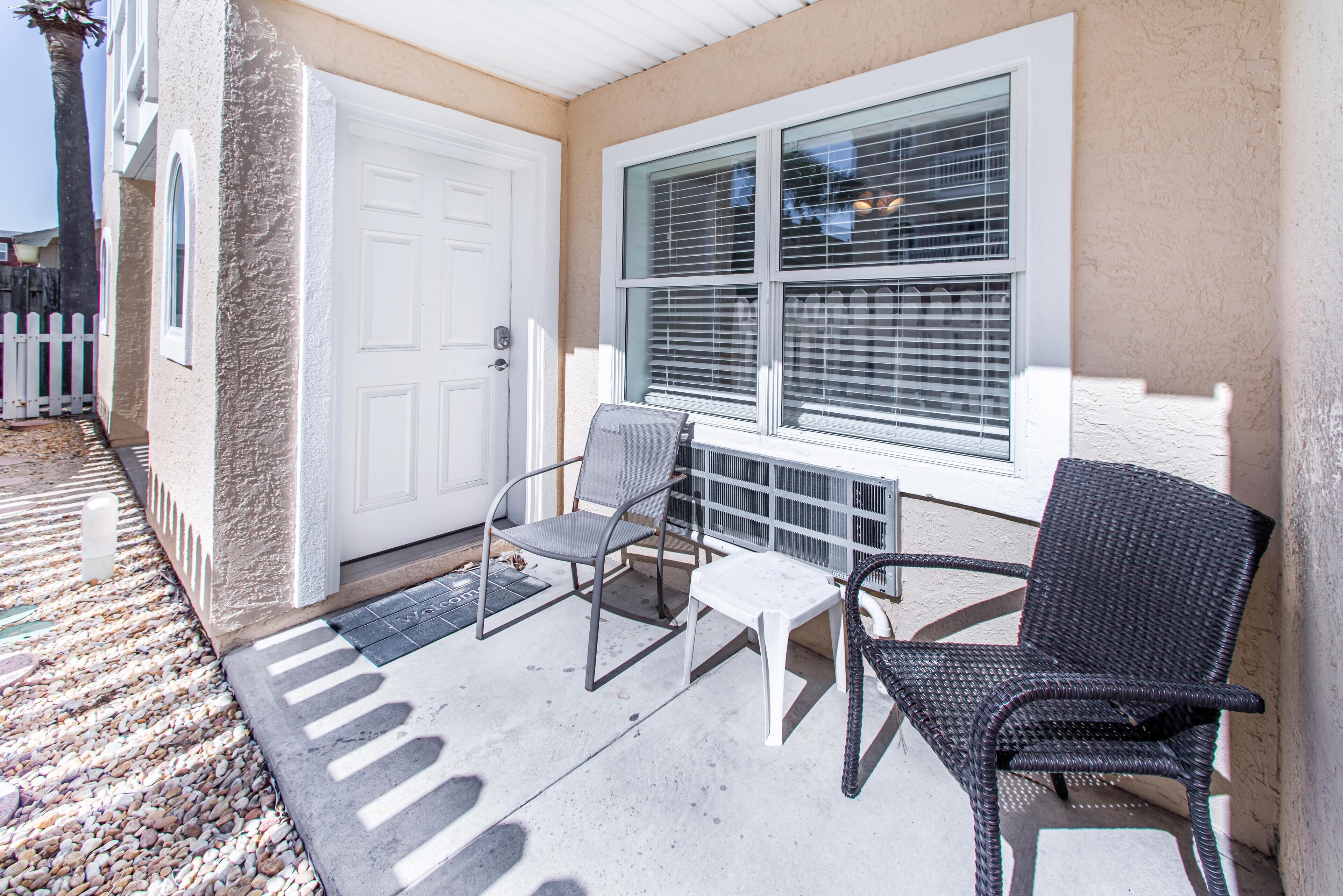 A covered patio with two chairs and a small table, a white door, and a window with blinds.