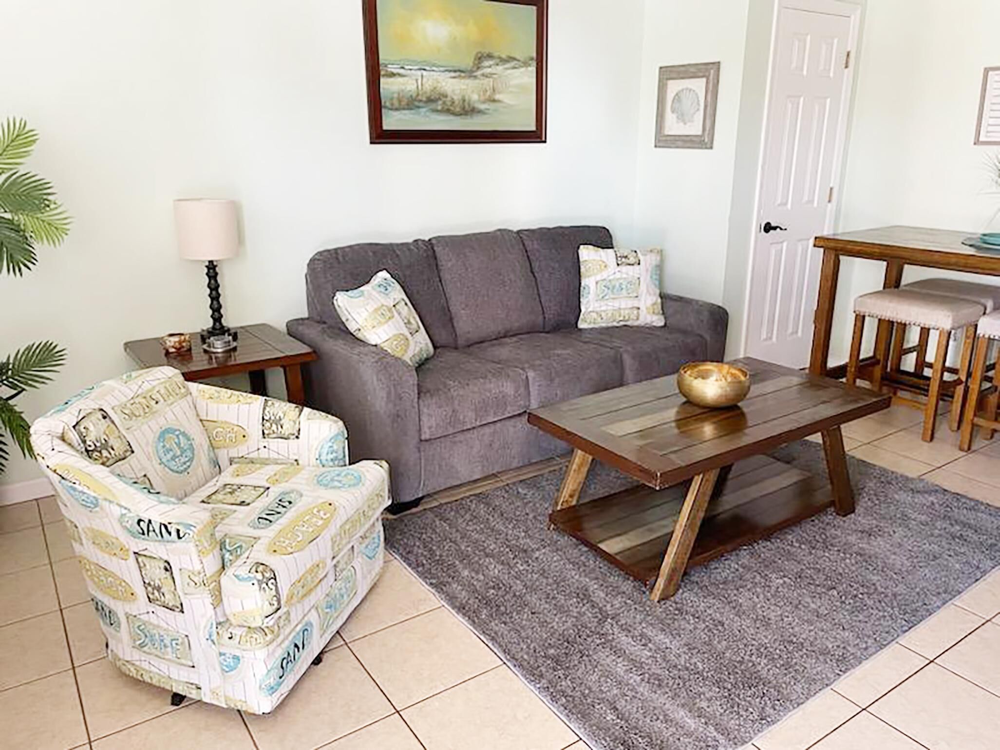Un salon avec un canapé gris, un fauteuil à motifs, une table basse en bois et une petite table à manger avec des tabourets.