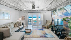 A living room with a sofa, coffee table, TV, and a view of the ocean through large windows. - Mariner West 205 (Panama City Beach)