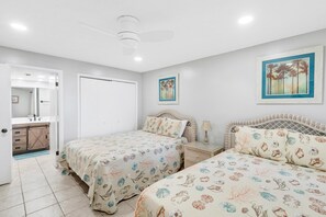 Quarto com duas camas, mesa de cabeceira, ventoinha no teto e porta para um banheiro.